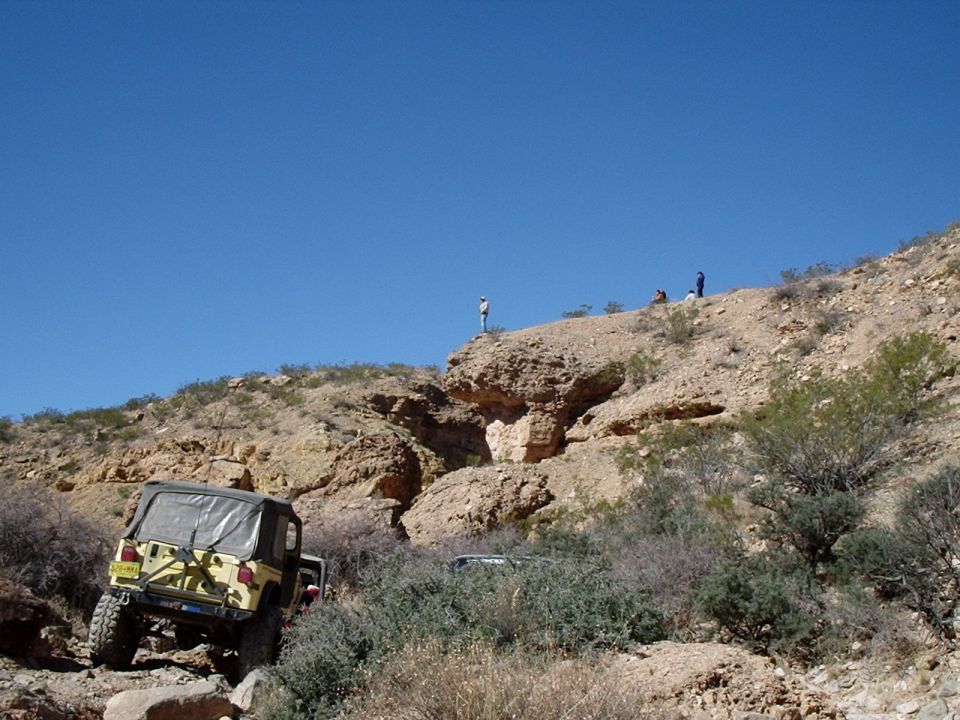 Yellow Jeep Wrangler climbing a rocky incline with other vehicles behind it on a sunny day