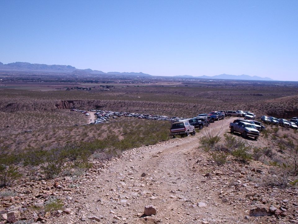 Dirt road with many parked vehicles, some in a line, others in a lot below. Mountains in the distance