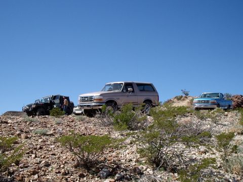 1996 Ford Bronco - A tan 1996 Ford Bronco is parked on a rocky hillside alongside two other off-road vehicles under a clear blue sky.