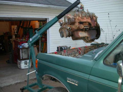 1990 Ford Bronco - Rusty V8 engine hoisted out of a green Ford Bronco on a green Carolina engine crane in a driveway, hanging above the empty engine bay with…