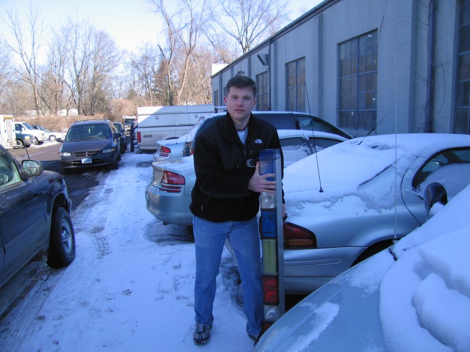 Man holding a car tail light assembly in a snowy parking lot with several cars