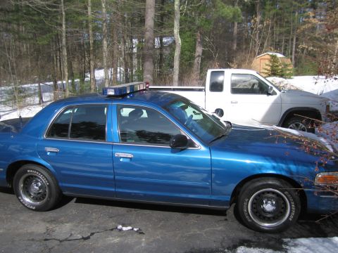 1991 Ford LTD Crown Victoria - A blue police sedan is parked next to a white pickup truck in a wooded area with snow on the ground.