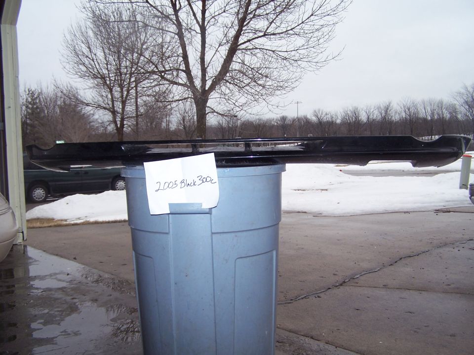Black car body part on gray trash can with '2005 Black 300c' sign outdoors in winter