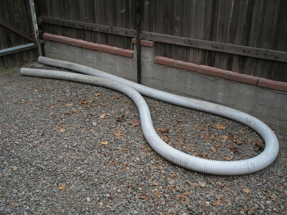 Two white flexible hoses lie on gravel in front of a wooden fence and concrete wall