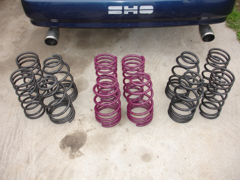 1993 Ford Taurus - Several sets of car springs are arranged on a concrete surface, with the rear of a blue SHO vehicle overhead.