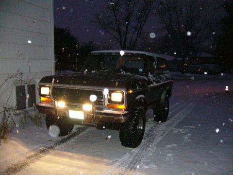 1979 Ford Bronco - A 1979 Ford Bronco is parked outside in the snow, its headlights illuminating the snowy driveway.
