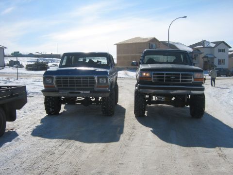 1995 Ford Bronco - Two lifted Ford trucks are parked side-by-side in a snowy area with houses in the background.