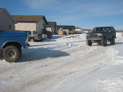 1995 Ford Bronco - A black Ford Bronco is being towed by a blue truck on a snowy street with houses in the background.