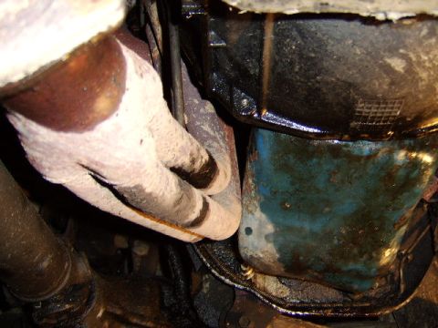 1989 Ford Bronco - Underside engine view showing an oil pan with visible oil residue and blue-green discoloration, alongside heat-wrapped exhaust piping insul…