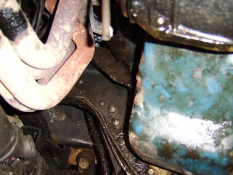 1989 Ford Bronco - Close-up underneath a vehicle showing greasy, oil-stained engine and transmission components, with a blue-painted metal pan or housing marr…