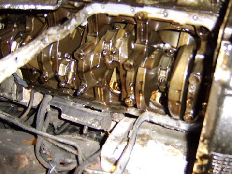 1989 Ford Bronco - Close-up of an oil-stained engine block bottom end with the oil pan removed, showing the crankshaft, main bearing caps and crankcase. Grime…