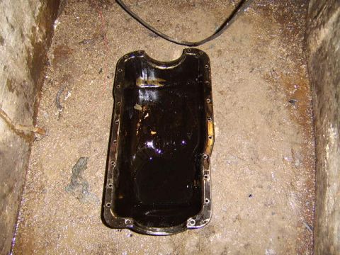 1989 Ford Bronco - A removed engine oil pan sits on a concrete floor, its interior coated with dark black oil and a bolt-hole flange around the rim. A black h…