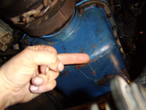 1989 Ford Bronco - A person's hand points at a blue engine oil pan showing surface rust, staining and possible seepage along the lower seam and gasket area. T…