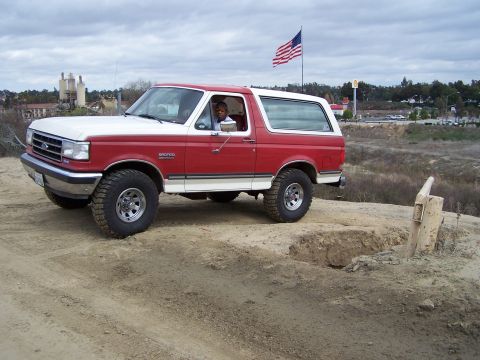 1989 Ford Bronco - A red and white Ford Bronco is parked on a dirt hill with an American flag in the background.