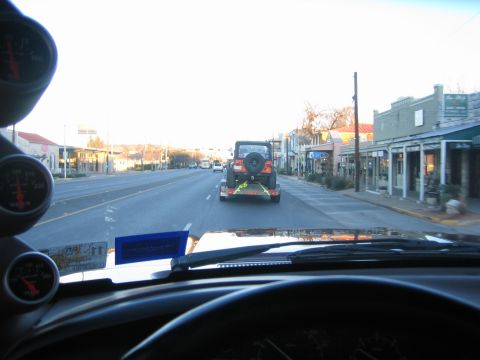 1996 Ford Bronco - A Jeep Wrangler is towed on a trailer down a street lined with shops.