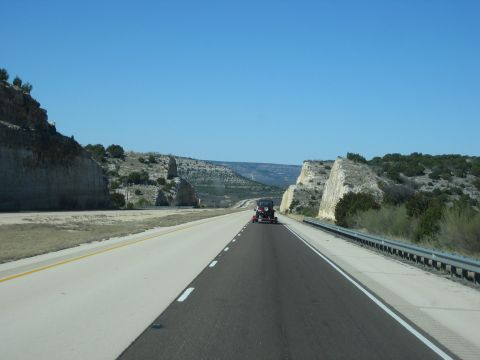 1996 Ford Bronco - A red SUV travels down a highway flanked by rocky cliffs under a clear blue sky.