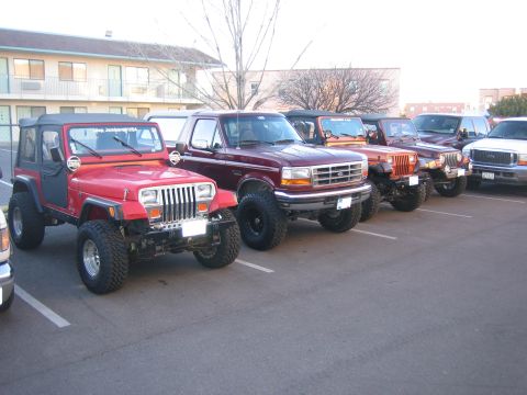 1996 Ford Bronco - A line of modified SUVs and trucks are parked in a lot, with a maroon Ford Bronco in the foreground.