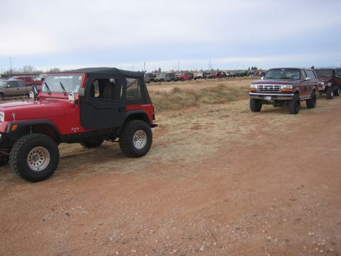 1996 Ford Bronco - A line of SUVs, including a maroon 1996 Ford Bronco, are parked on a dirt lot under a cloudy sky.