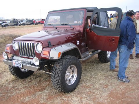 1996 Ford Bronco - A maroon Jeep Wrangler is parked on dry grass with its driver's door open, and several other vehicles are visible in the background.