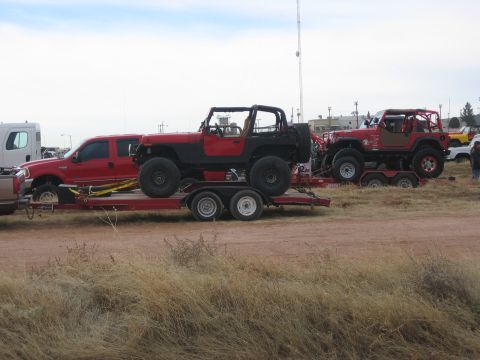 1996 Ford Bronco - Two red Jeeps are transported on trailers, one behind a red truck, under a cloudy sky.