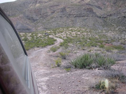 1996 Ford Bronco - Two SUVs navigate a dusty, winding road through a sparse desert terrain with rocky hills in the background.