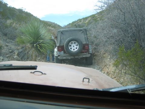 1996 Ford Bronco - Two Jeeps navigate a narrow, rocky trail through a desert landscape, with one vehicle in the foreground and another ahead.
