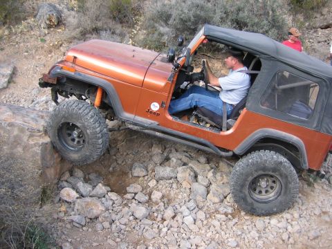 1996 Ford Bronco - An orange Jeep Wrangler is shown off-roading on a rocky trail with a driver at the wheel.