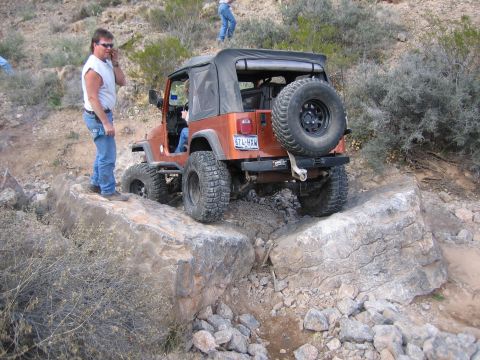 1996 Ford Bronco - An orange Jeep Wrangler is shown off-roading over large boulders on a dry, rocky landscape.