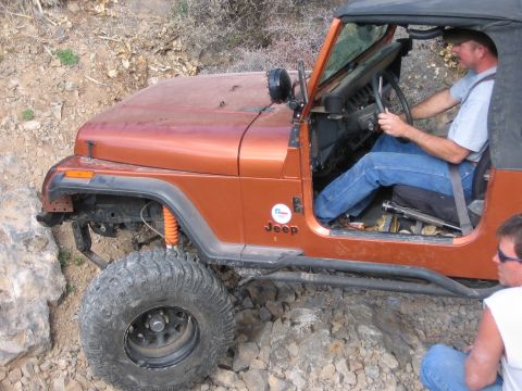 1996 Ford Bronco - A man drives an orange Jeep Wrangler off-road, navigating a rocky path with a passenger looking on.