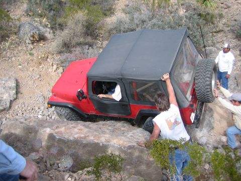 1996 Ford Bronco - A red Jeep Wrangler struggles over large rocks on an off-road trail, with several individuals helping to guide it.