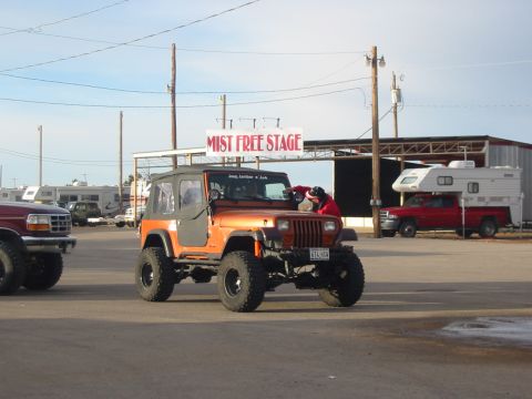 1996 Ford Bronco - Orange Jeep Wrangler parked near a "Mist Free Stage" sign, with other vehicles in the background.