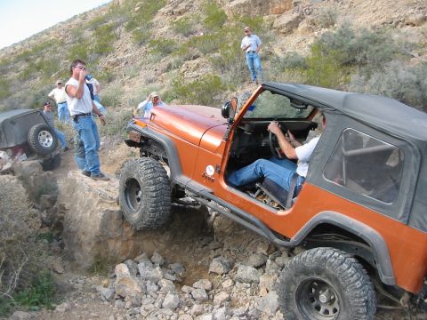 1996 Ford Bronco - A group of people watch as an orange Jeep Wrangler tackles a challenging off-road course.