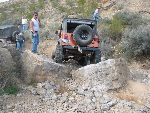 1996 Ford Bronco - An orange Jeep Wrangler maneuvers over large rocks on an off-road trail, with another vehicle and people visible in the background.