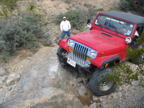 1996 Ford Bronco - A red Jeep Wrangler tackles a challenging rocky path, with a "Jeep Jamboree USA" sticker visible on the windshield.