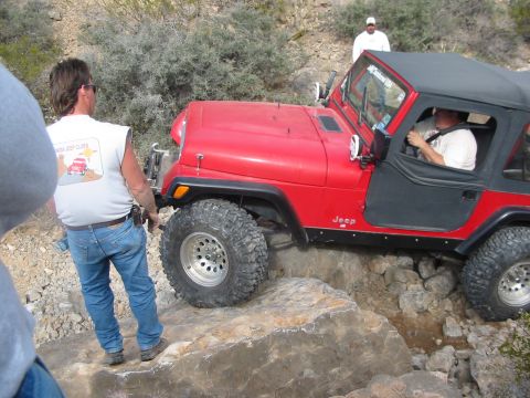 1996 Ford Bronco - A red Jeep Wrangler is shown off-roading on a rocky terrain, with two people present.