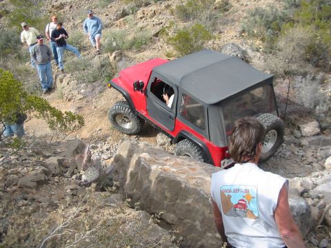 1996 Ford Bronco - A red Jeep Wrangler is shown off-roading on a steep, rocky incline with spectators present.