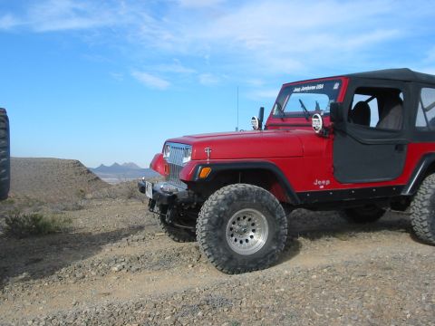 1996 Ford Bronco - A red Jeep Wrangler is parked on a rocky incline under a blue sky, with a "Jeep Jamboree USA" sticker visible on the windshield.