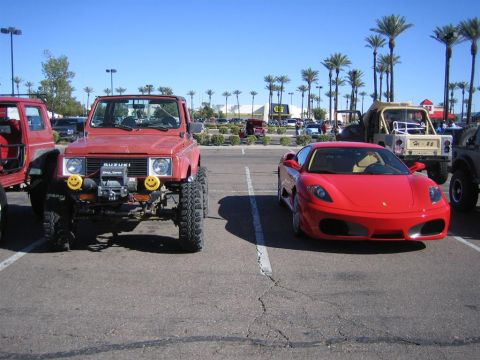 1987 Suzuki Samurai - A modified 1987 Suzuki Samurai with oversized tires and a winch sits parked next to a red Ferrari in a parking lot.