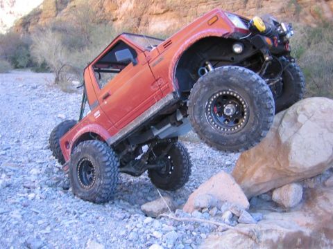 1987 Suzuki Samurai - An orange 1987 Suzuki Samurai navigates a rocky terrain with its front right wheel high in the air.