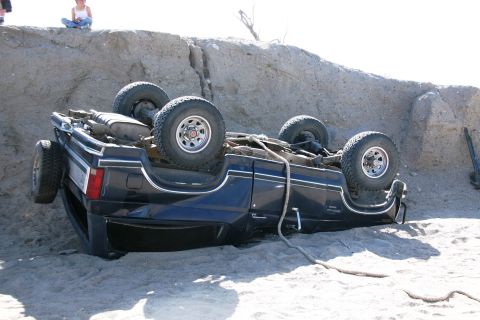 1990 Ford Bronco - dark blue pickup truck rests overturned on its roof in the sand against a dune embankment, wheels in the air.
