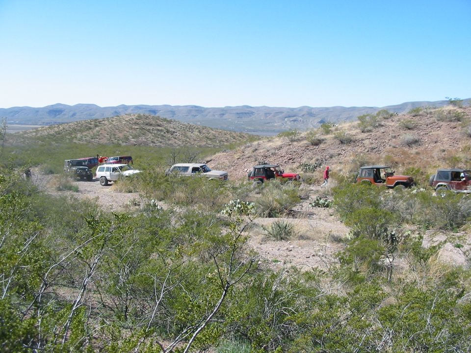 Several off-road vehicles driving on a dirt trail through a desert landscape with hills in the background