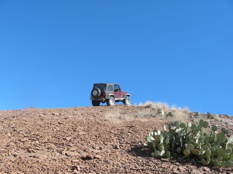 1996 Ford Bronco - A maroon Jeep Wrangler is parked on a rocky incline, with prickly pear cactus in the foreground and a vast blue sky above.