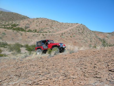 1996 Ford Bronco - A red Jeep Wrangler navigates a , dry terrain, ascending a dusty trail on a barren hill.