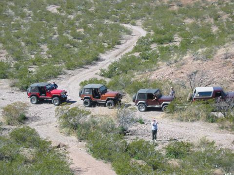 1996 Ford Bronco - Several Jeeps are stopped on a dirt path in a dry, arid landscape with low-lying bushes.