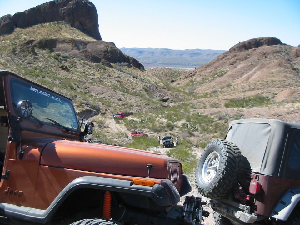 Several Jeeps driving on a rocky desert trail with mountains in the background. One orange Jeep is in the foreground