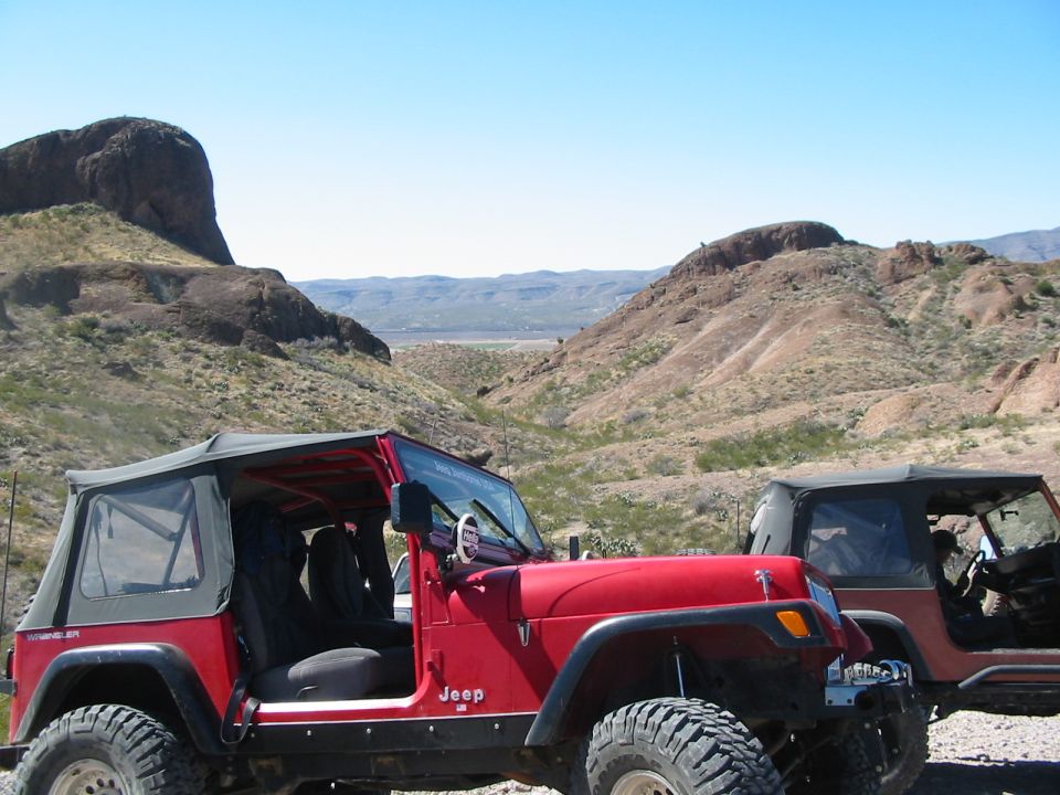 Red Jeep Wrangler with soft top, off-road tires, and a second Jeep behind it in a desert landscape