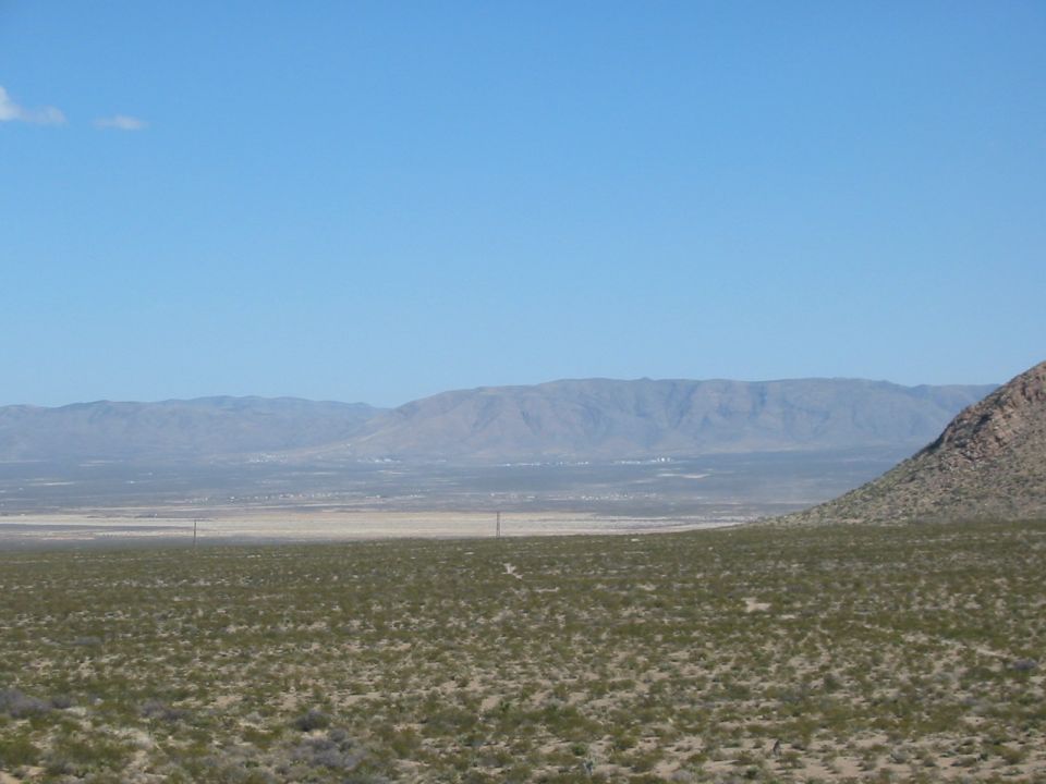 wide view of a desert landscape with scrub brush in the foreground, flat plains in the midground, and mountains in the background under a clear blue sky