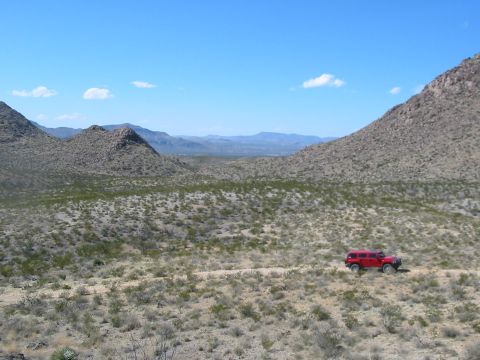 1996 Ford Bronco - A red SUV drives along a dirt path through a barren, mountainous desert under a clear blue sky.