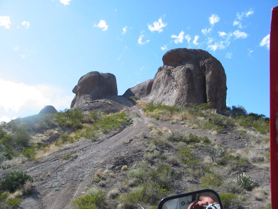 Desert landscape with large rock formations and sparse vegetation under a blue sky with clouds. A side mirror with a reflection of a person holding a camera is visible in the foreground