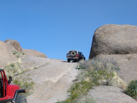 1996 Ford Bronco - Two off-road vehicles, a red Jeep and a 1996 Ford Bronco, ascend a steep dirt trail under a clear blue sky.